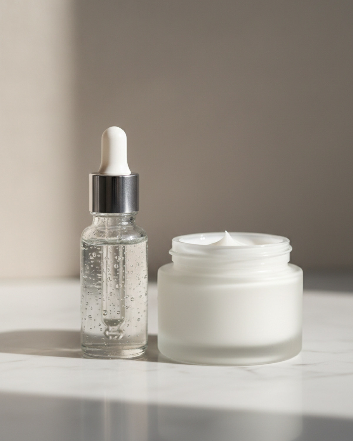 A glass dropper bottle of clear serum and a jar of cream on a pale marble counter in soft morning light